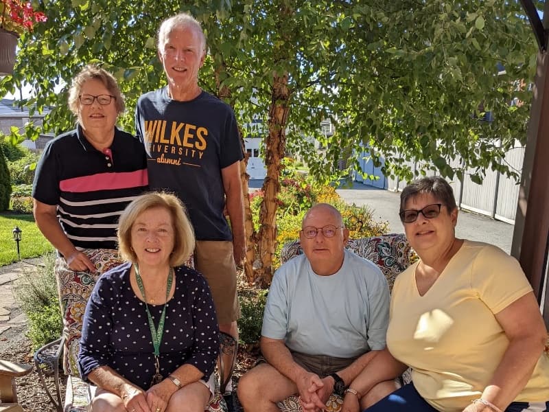 A group of five people poses together outdoors in a garden setting. A group of five people poses together outdoors in a garden setting.
