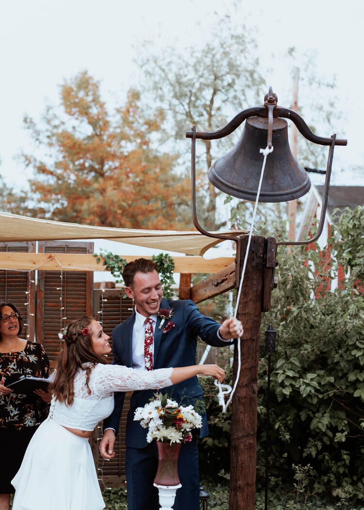 A bride and groom joyfully ring a large bell during their wedding ceremony, with guests looking on.