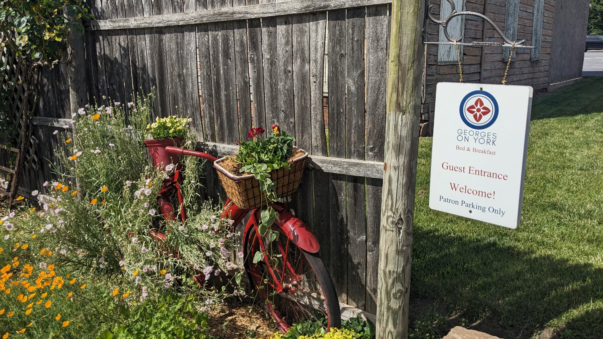 A red bicycle adorned with flower baskets is positioned next to a wooden fence and a sign for the guest entrance.