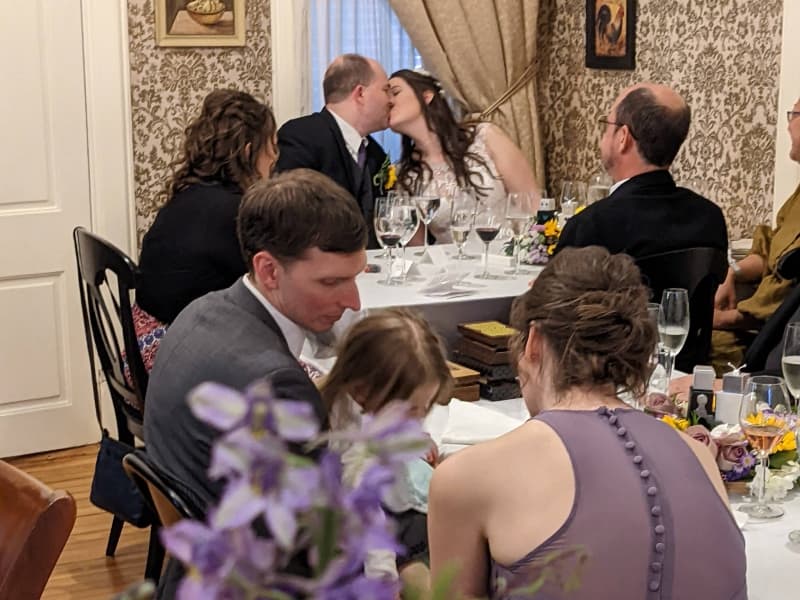 A couple shares a kiss while seated at a wedding reception table surrounded by guests. A couple shares a kiss while seated at a wedding reception table surrounded by guests.
