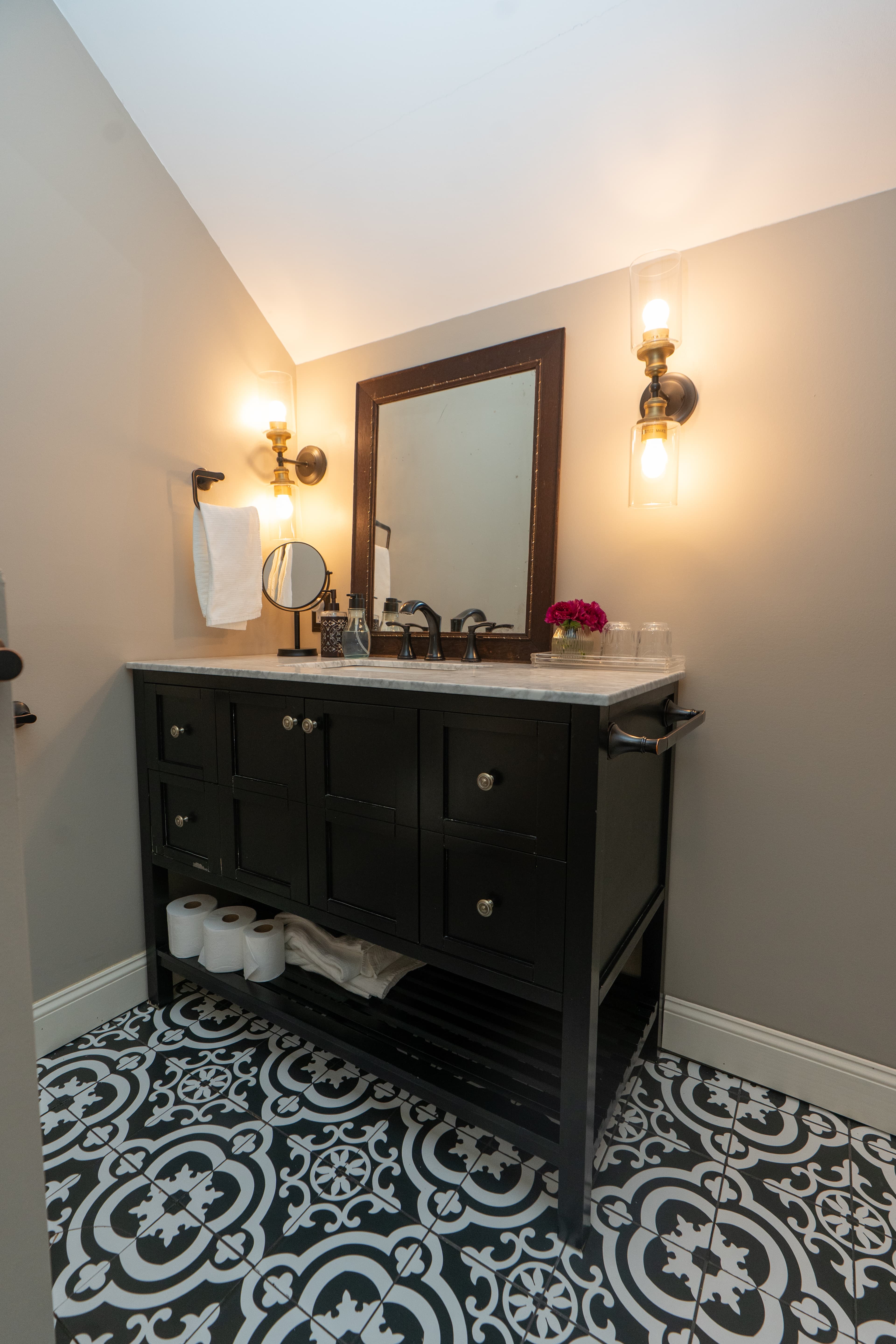 A high-angle view of a bathroom vanity with a dark wood cabinet, a white marble countertop, and a square white sink, featuring a large framed mirror flanked by two elegant wall sconces.